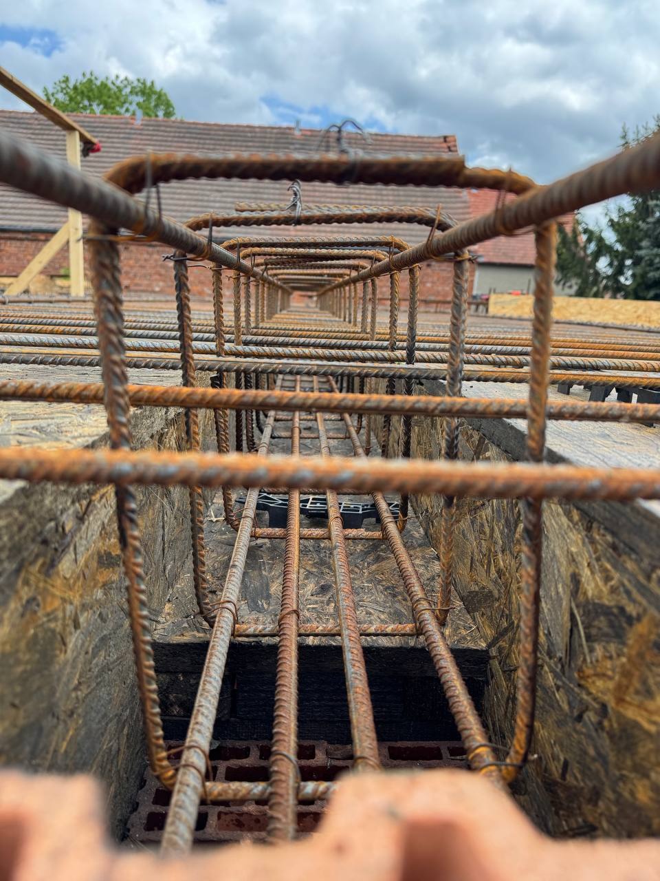Zbrojenie fundamentów w wykopie, widok z dołu. Stalowe pręty tworzą siatkę, na tle cegieł i desek. Niebo z chmurami w tle.
