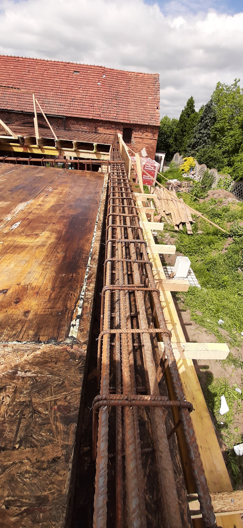 Zbrojenie pionowe w szalunku pod ścianę fundamentową na budowie domu jednorodzinnego. Widoczne drewniane deski szalunkowe i elementy konstrukcyjne.