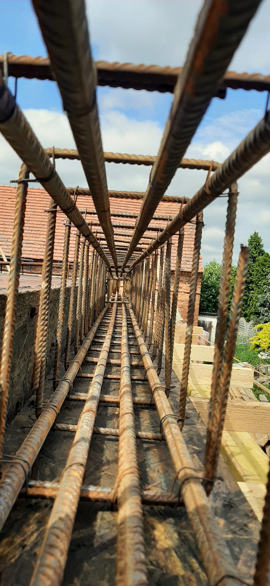 Zbrojenie fundamentów domu jednorodzinnego, widok z dołu na konstrukcję z prętów stalowych, w tle fragment budynku z czerwoną dachówką i zielenią.