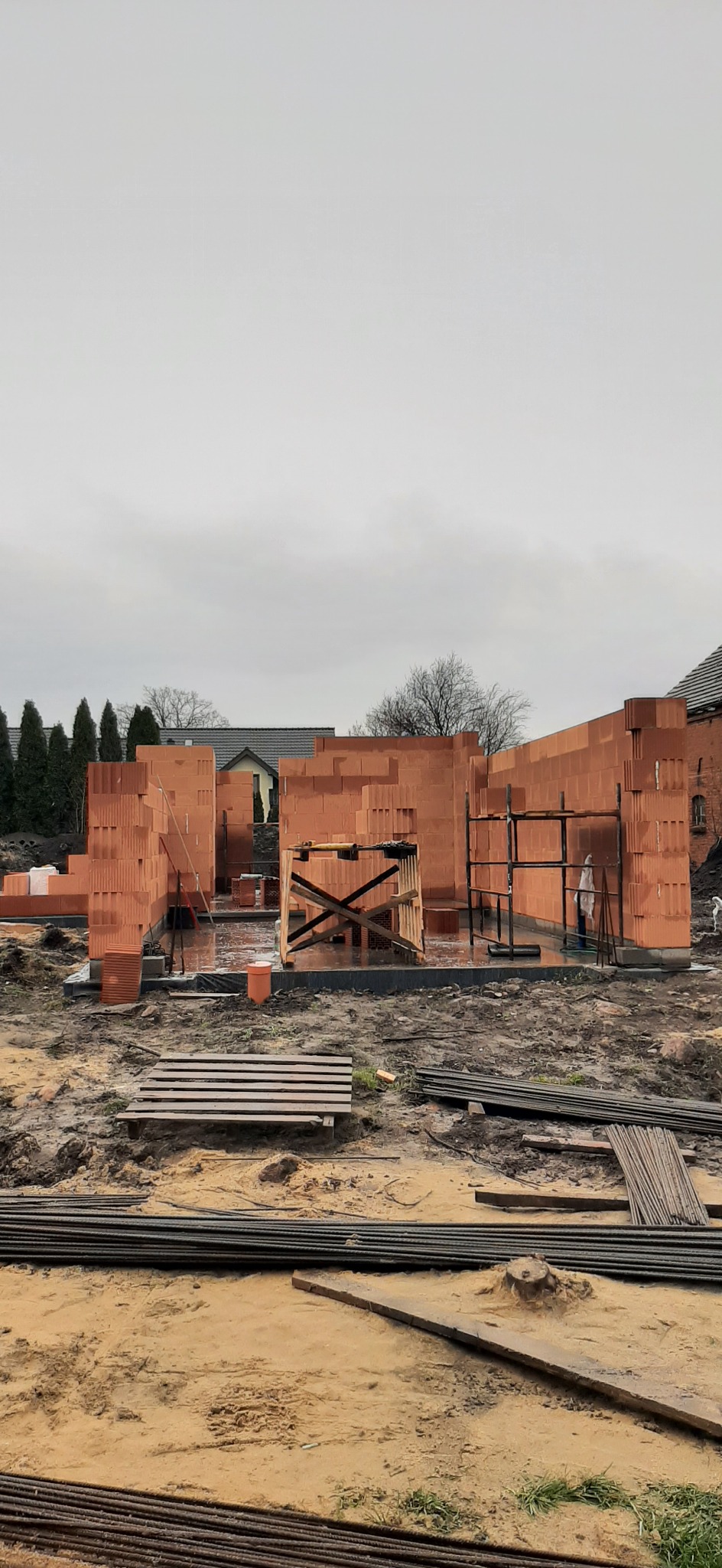 Wznoszenie ścian z czerwonej ceramiki poryzowanej na budowie domu. Widoczne rusztowania i przygotowane materiały. Pochmurne niebo w tle.