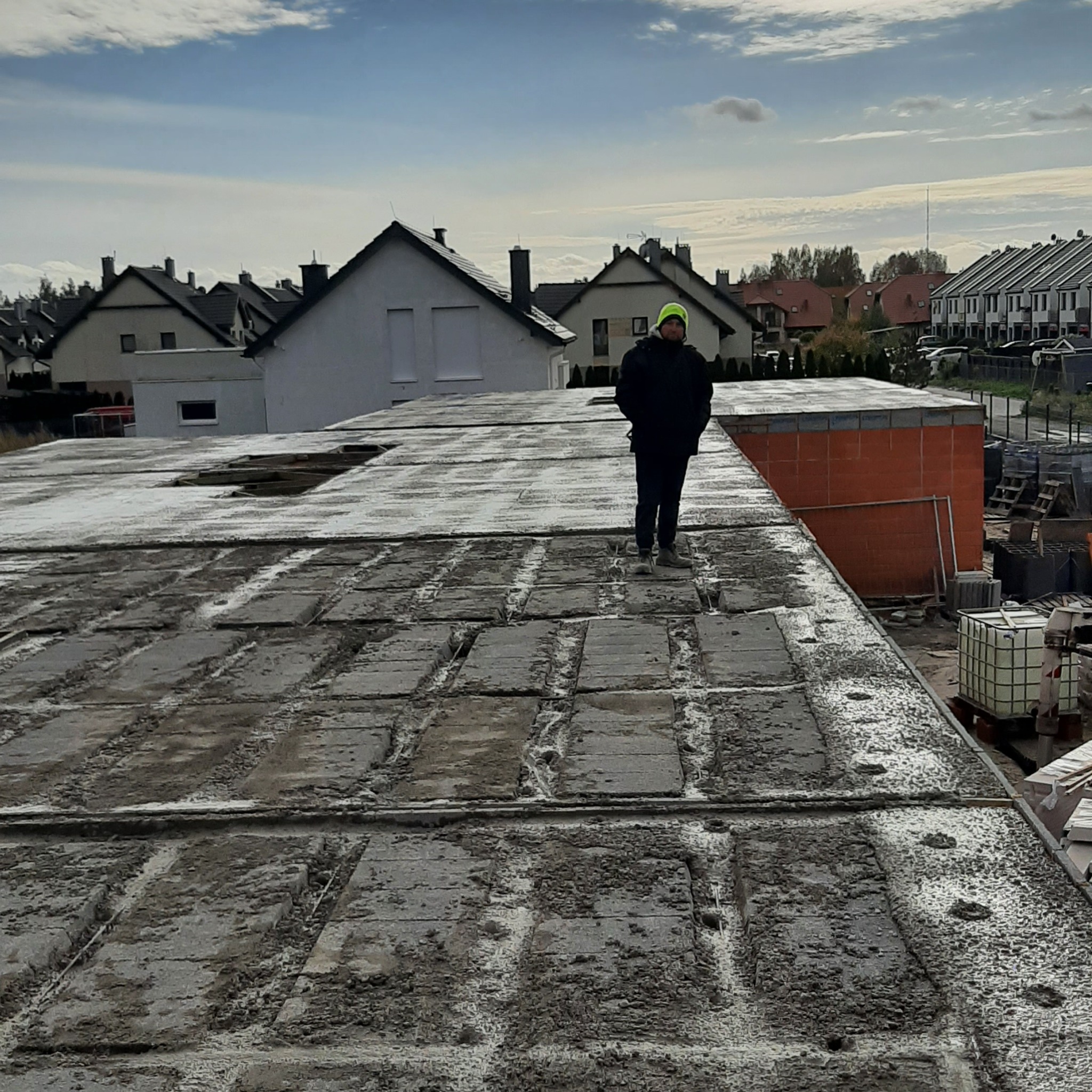Budowa domu: mężczyzna w neonowej czapce stoi na betonowym stropie, w tle widać budynki mieszkalne i częściowo wymurowaną ścianę z czerwonej cegły.