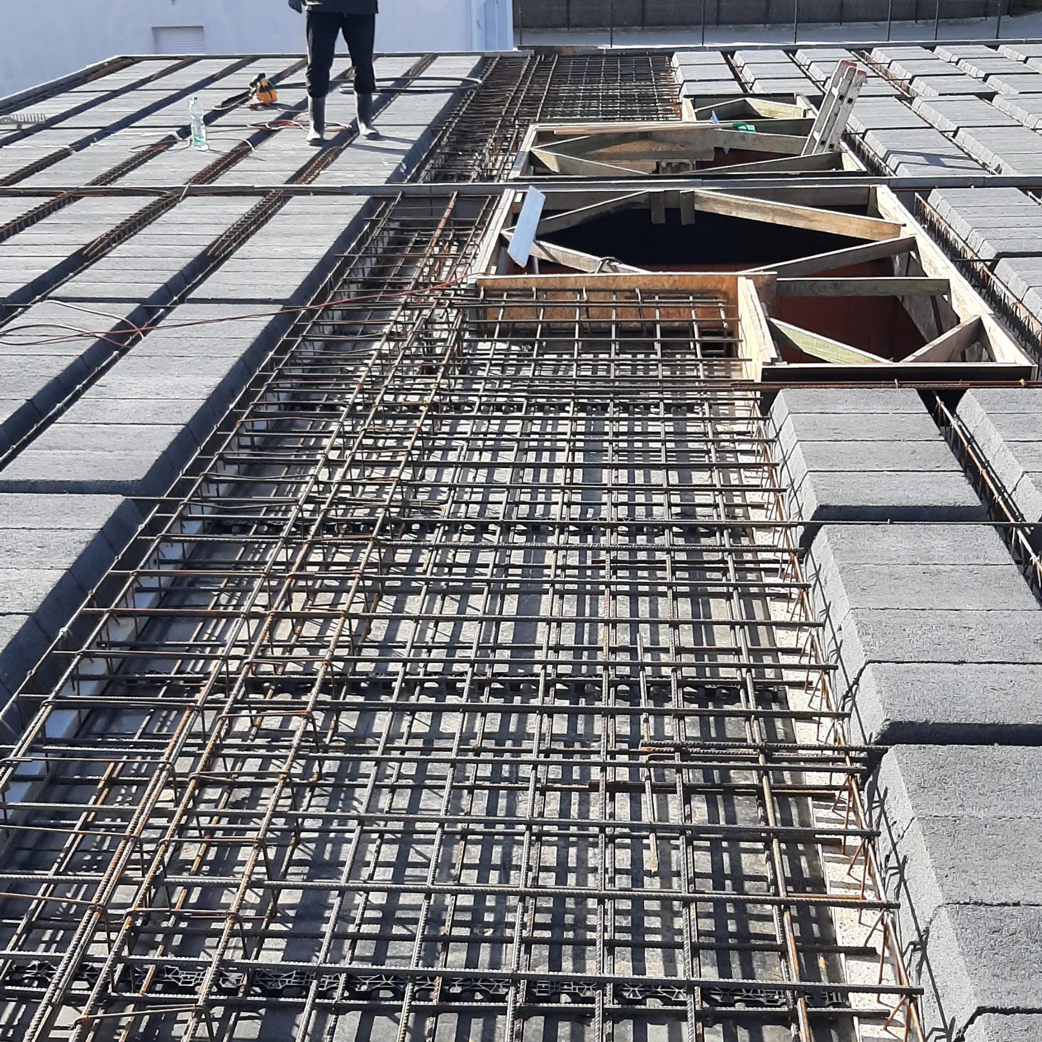 Zbrojenie stropu Teriva z szalunkiem i pustakami betonowymi, widok z góry. Na górze kadru widoczna osoba w odzieży roboczej i narzędzia.