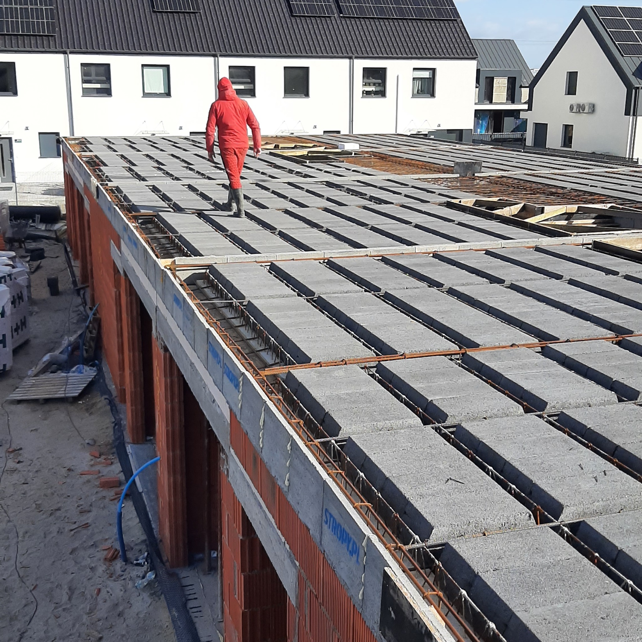 Budowa domu: strop Teriva zbrojony stalą, robotnik w czerwonym kombinezonie na tle białych domów z panelami słonecznymi. Widok z góry na plac budowy.