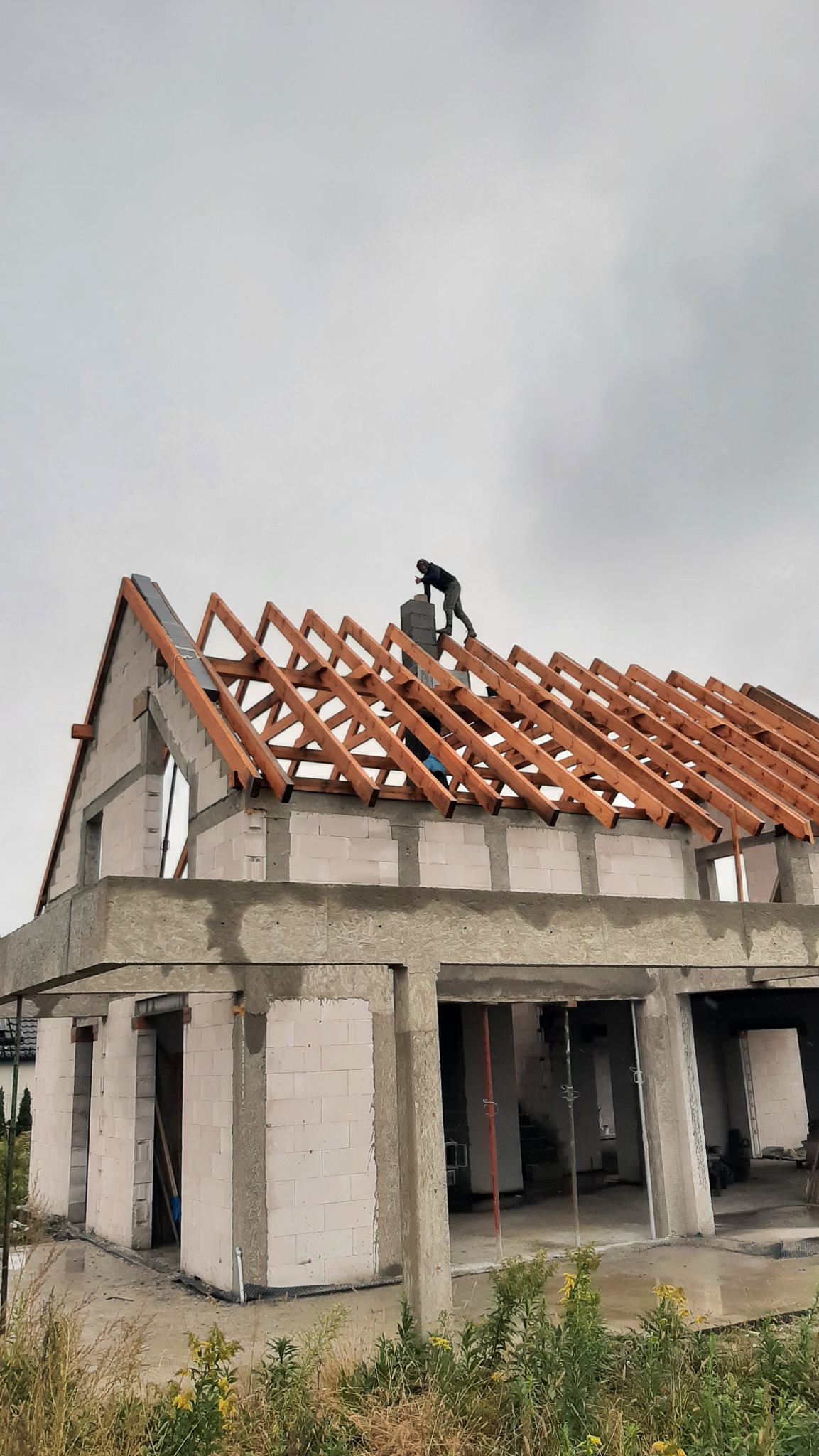 Budowa domu w trakcie realizacji: murowane ściany z betonu komórkowego, drewniana konstrukcja dachu i pracownik na dachu, pochmurne niebo w tle.