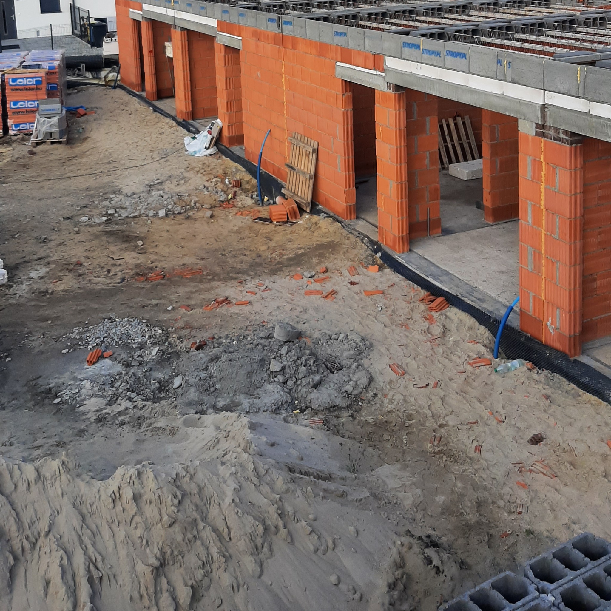 Budowa bliźniaka z czerwonej cegły. Widoczne ściany nośne, nadproża i strop. Na ziemi gruz i piasek. Paleta z cegłami Leier w tle.