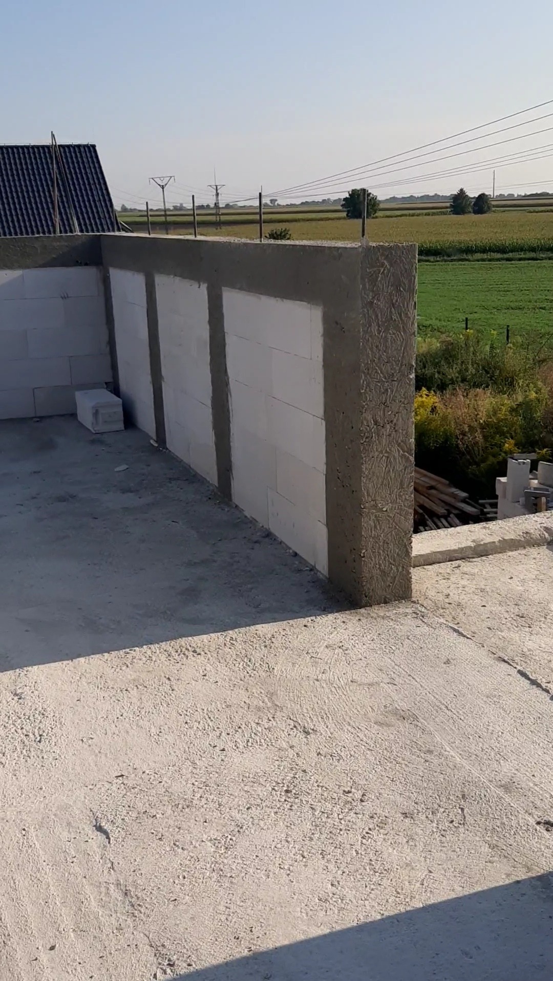 Ściana z betonu komórkowego w trakcie murowania, widok z góry na plac budowy z polami w tle. Świeżo nałożona zaprawa na pionowych elementach.