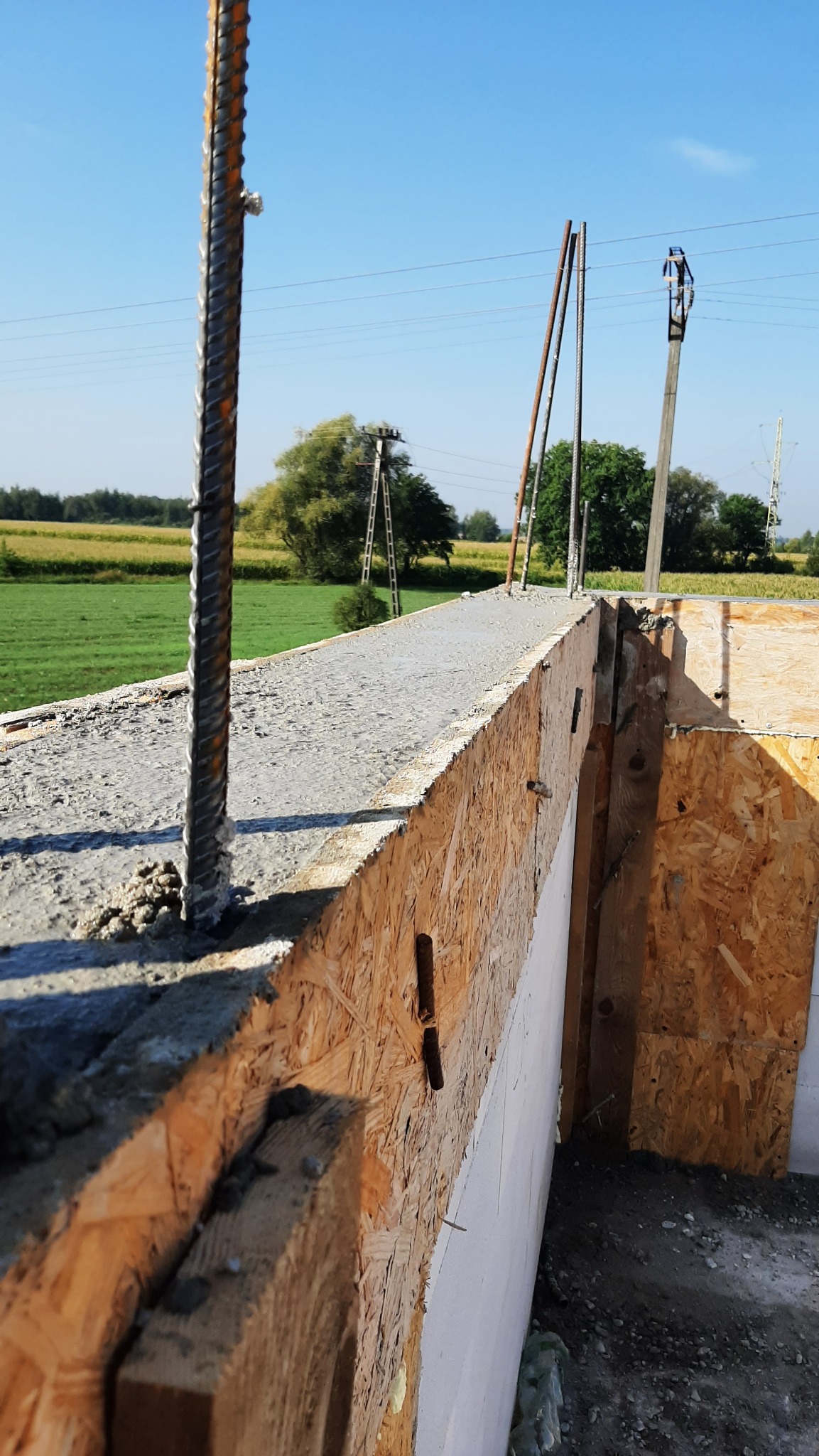 Zalewanie betonem szalunku zbrojonego na fundamencie domu jednorodzinnego. Widok na konstrukcję zbrojoną, szalunek z płyty OSB i piankę uszczelniającą. W tle zielone pola i słupy energetyczne.