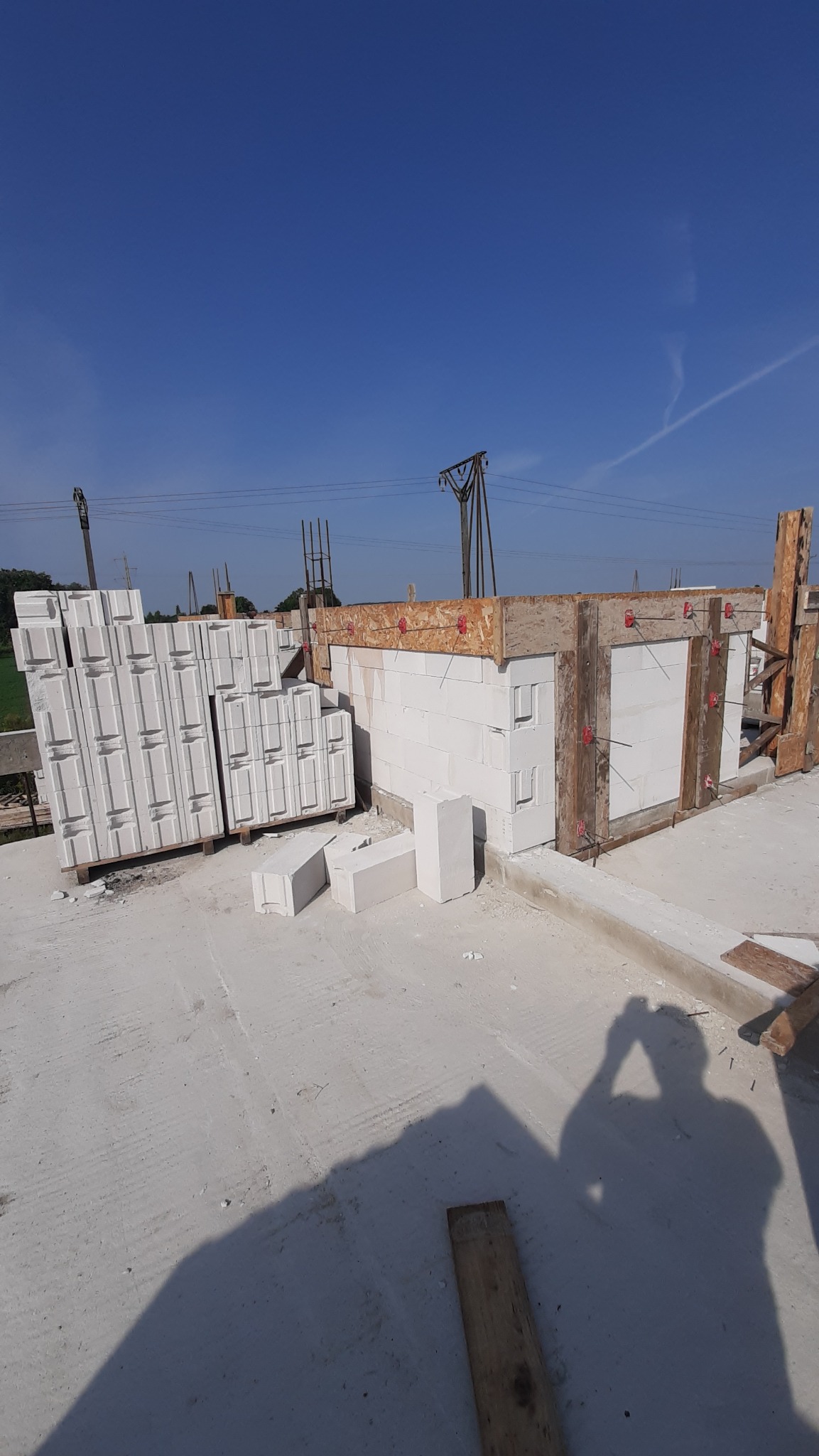 Wznoszenie ścian z białych bloczków na budowie domu, widoczne szalunki i zapas materiału. Na pierwszym planie cień osoby robiącej zdjęcie, błękitne niebo w tle.