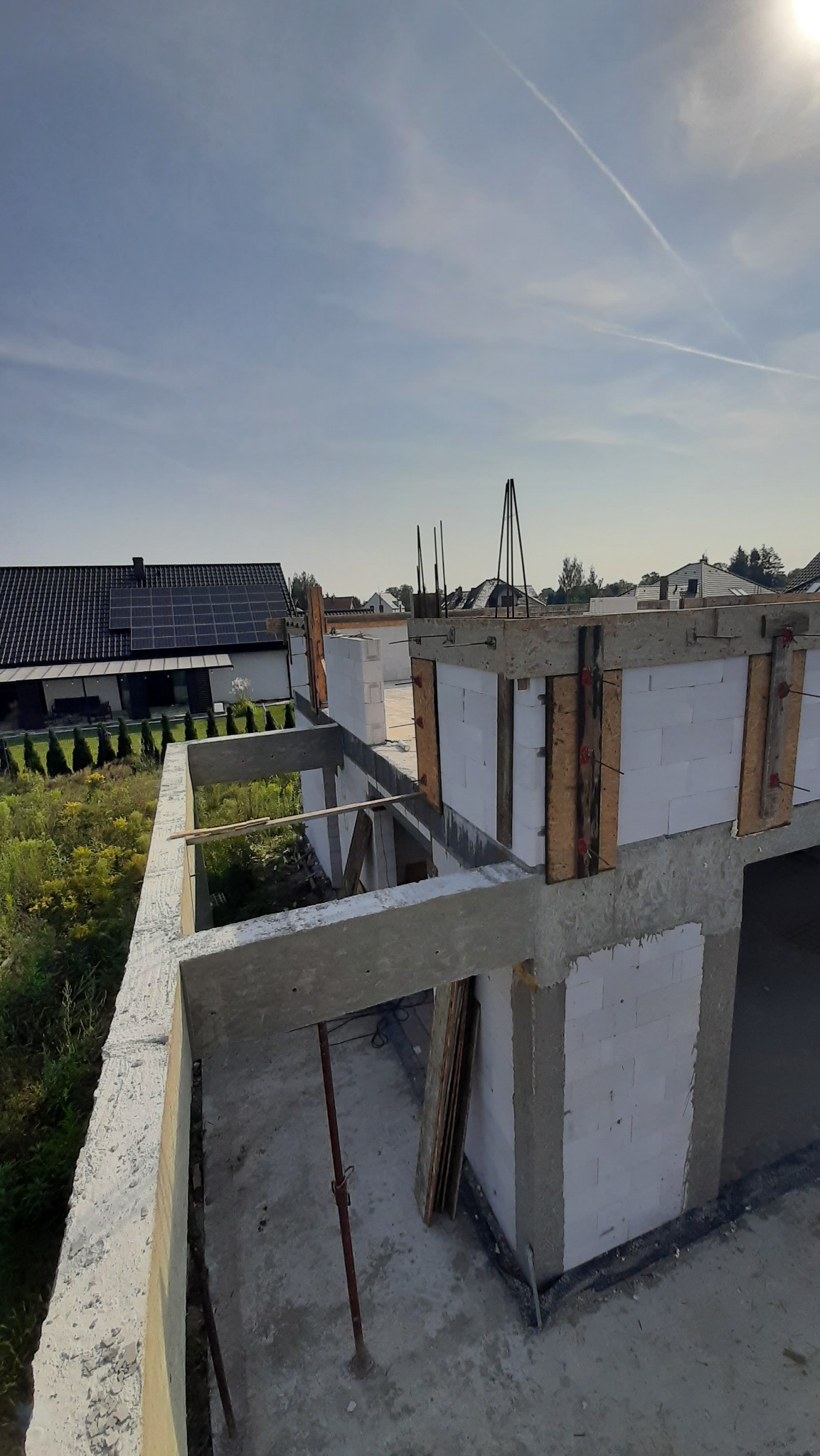 Wznoszenie ścian domu z betonu komórkowego, widoczne szalunki i zbrojenia. W tle dom jednorodzinny z panelami słonecznymi. Ujęcie z góry.