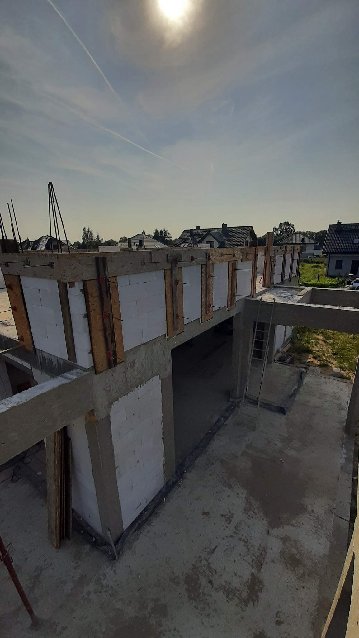 Wysoki kąt na budowę domu z betonu komórkowego: ściany w szalunkach, zbrojenie, widok na okoliczne domy w słoneczny dzień, drabina oparta o konstrukcję.