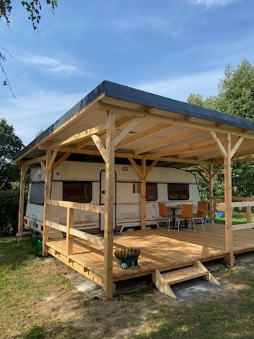 Drewniana konstrukcja z zadaszeniem nad przyczepą kempingową, z tarasem i schodami, otoczona zielenią.