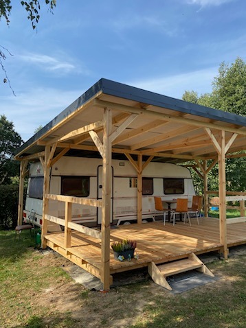 Zadaszony taras z drewna przyczepy kempingowej z drewnianą balustradą, schodkami i doniczką z wrzosami, na trawniku w słoneczny dzień.