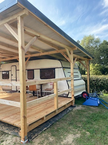 Zadaszona, drewniana konstrukcja nad tarasem z jasnego drewna, obok zaparkowana przyczepa kempingowa, widoczny wózek budowlany z niebieską skrzynią.