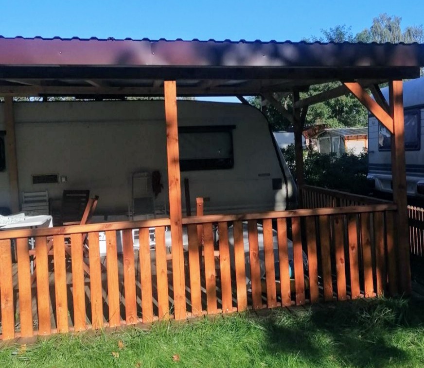 Zadaszony taras drewniany z niską balustradą, przylegający do przyczepy kempingowej, widok z zewnątrz w słoneczny dzień.