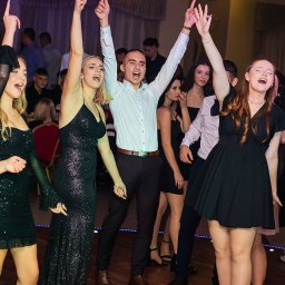 Fotografia Cichowski - Grupa rozbawionych młodych ludzi w eleganckich strojach tańczy i śpiewa na parkiecie podczas imprezy, z uniesionymi rękami i radosnymi minami.