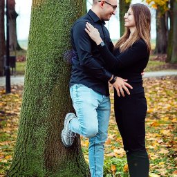 Fotografia Cichowski - Para zakochanych oparta o drzewo w jesiennym parku, on w czarnej koszuli i jasnych jeansach, ona w czarnym stroju, oboje w sportowych butach i okularach.