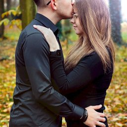 Fotografia Cichowski - Mężczyzna w okularach całuje kobietę w czoło na tle jesiennego lasu, oboje ubrani na czarno, ona ma długie blond włosy.