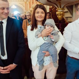 Fotografia Cichowski - Uśmiechnięta matka trzyma na rękach elegancko ubranego chłopca w szarej czapeczce, obok mężczyźni w kościele, jeden trzyma świecę