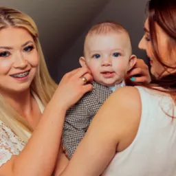 Dwie kobiety trzymające niemowlę w eleganckim ubranku; blondynka z aparatem ortodontycznym i pierścionkiem na palcu, uśmiechnięte dziecko z niebieskimi oczami.