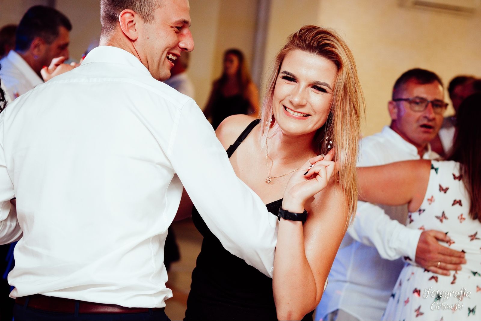 Uśmiechnięta blondynka w czarnej sukience tańczy z mężczyzną w białej koszuli na parkiecie podczas imprezy, w tle widoczni inni goście.