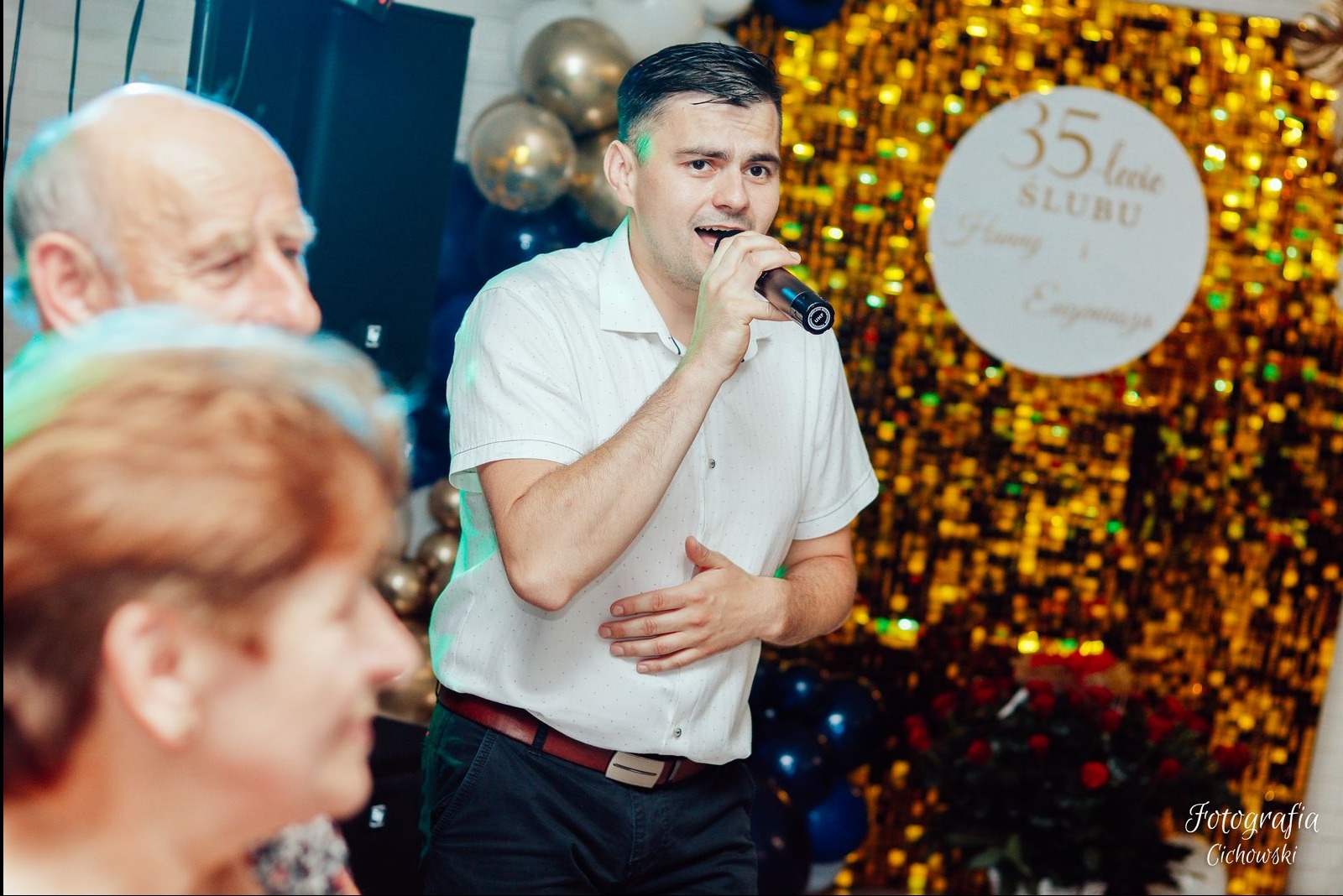 Mężczyzna w białej koszuli śpiewa do mikrofonu na tle dekoracji z okazji 35-lecia ślubu, widoczni goście w tle.