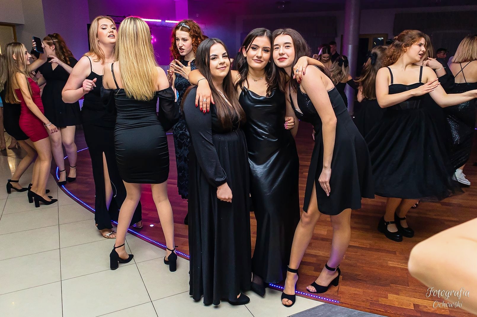Grupa młodych kobiet w czarnych sukienkach pozuje do zdjęcia podczas imprezy, parkiet z podświetleniem LED, widoczne osoby w tle.