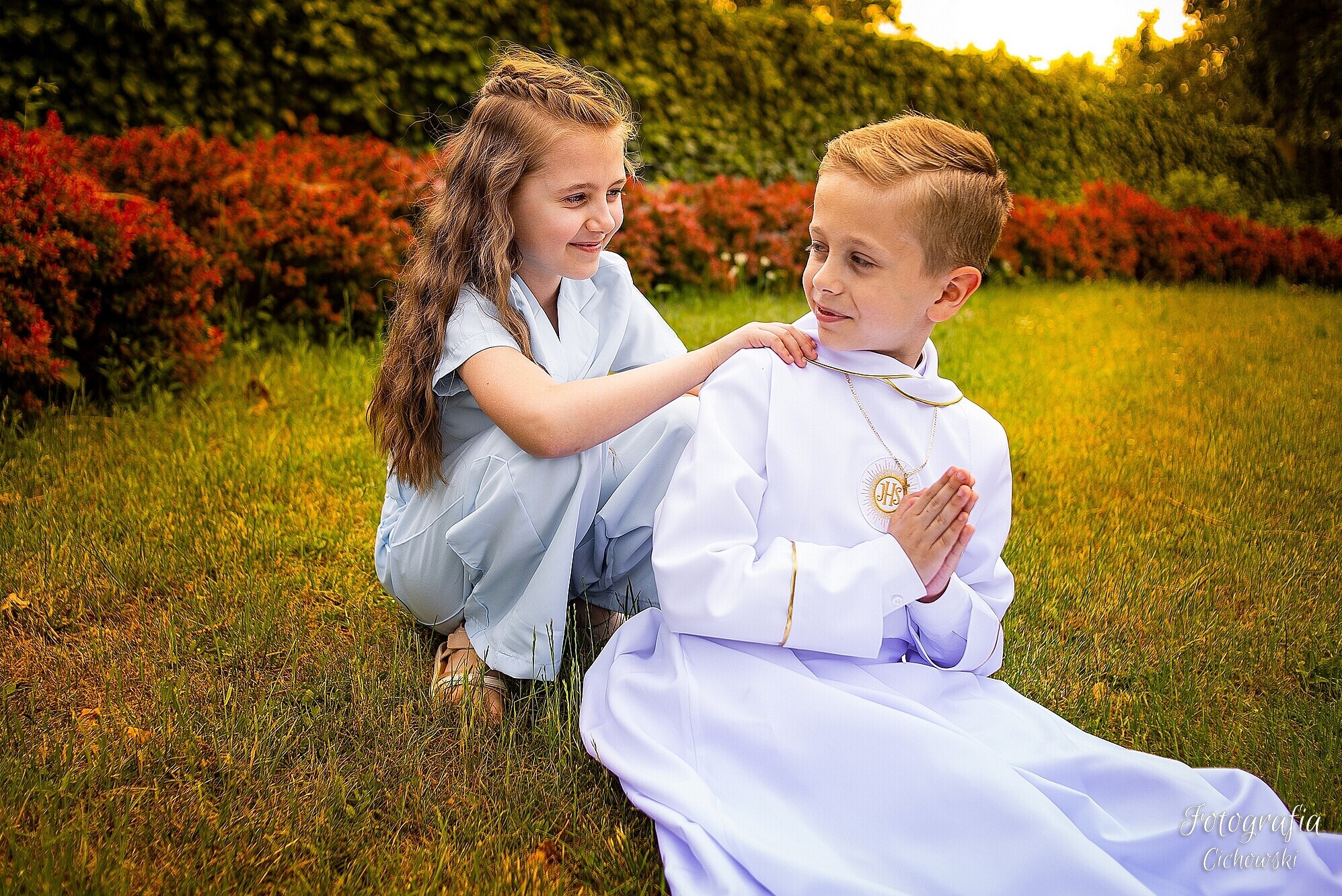 Uśmiechnięta dziewczynka w jasnoniebieskim stroju kuca obok chłopca w białej albie, który składa dłonie do modlitwy, złoty medalion na szyi, tło z zielonej trawy i czerwonych krzewów.