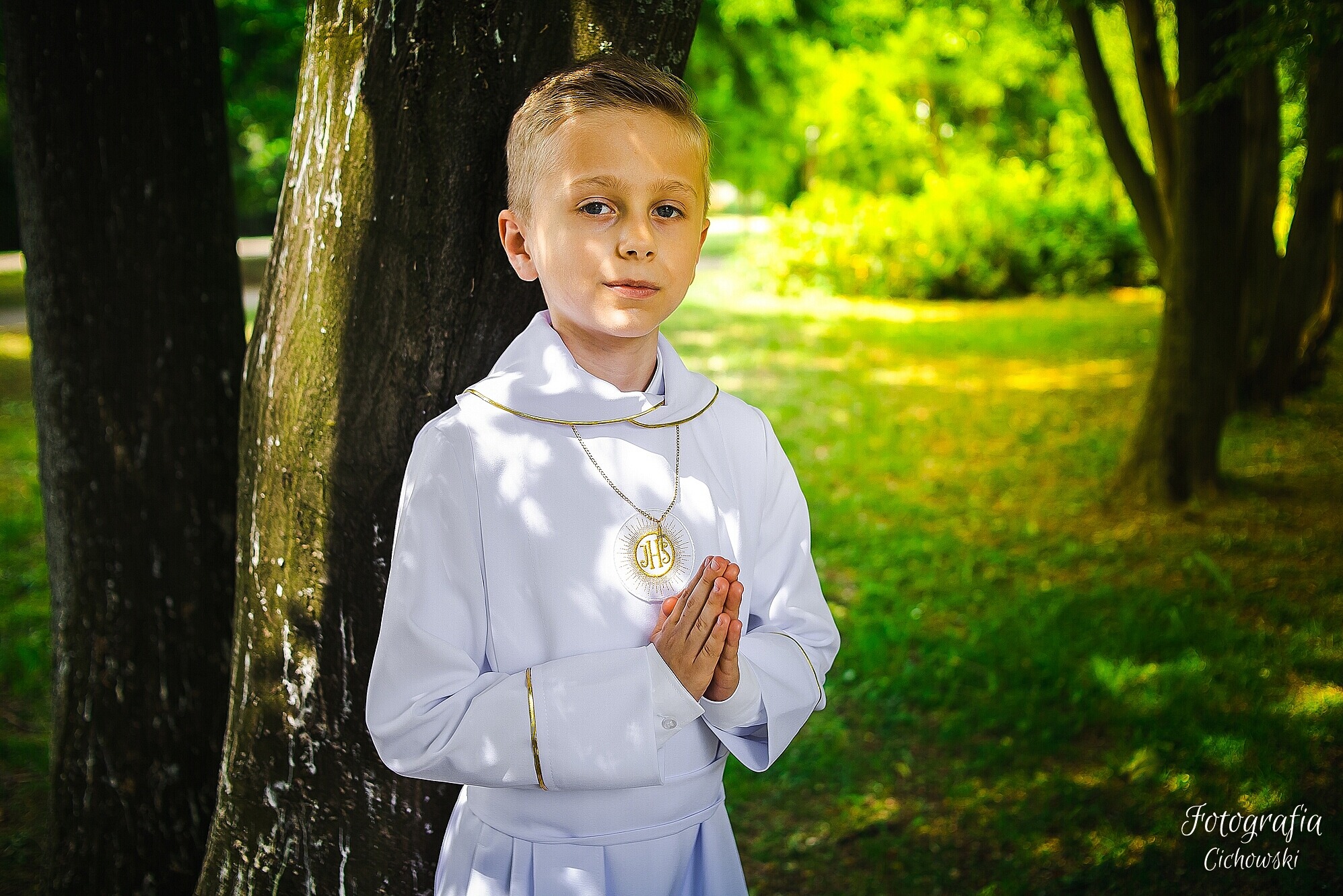 Chłopiec w stroju komunijnym, oparty o drzewo w parku, z złączonymi dłońmi i medalikiem IHS na szyi, w kontraście z zielonym tłem.