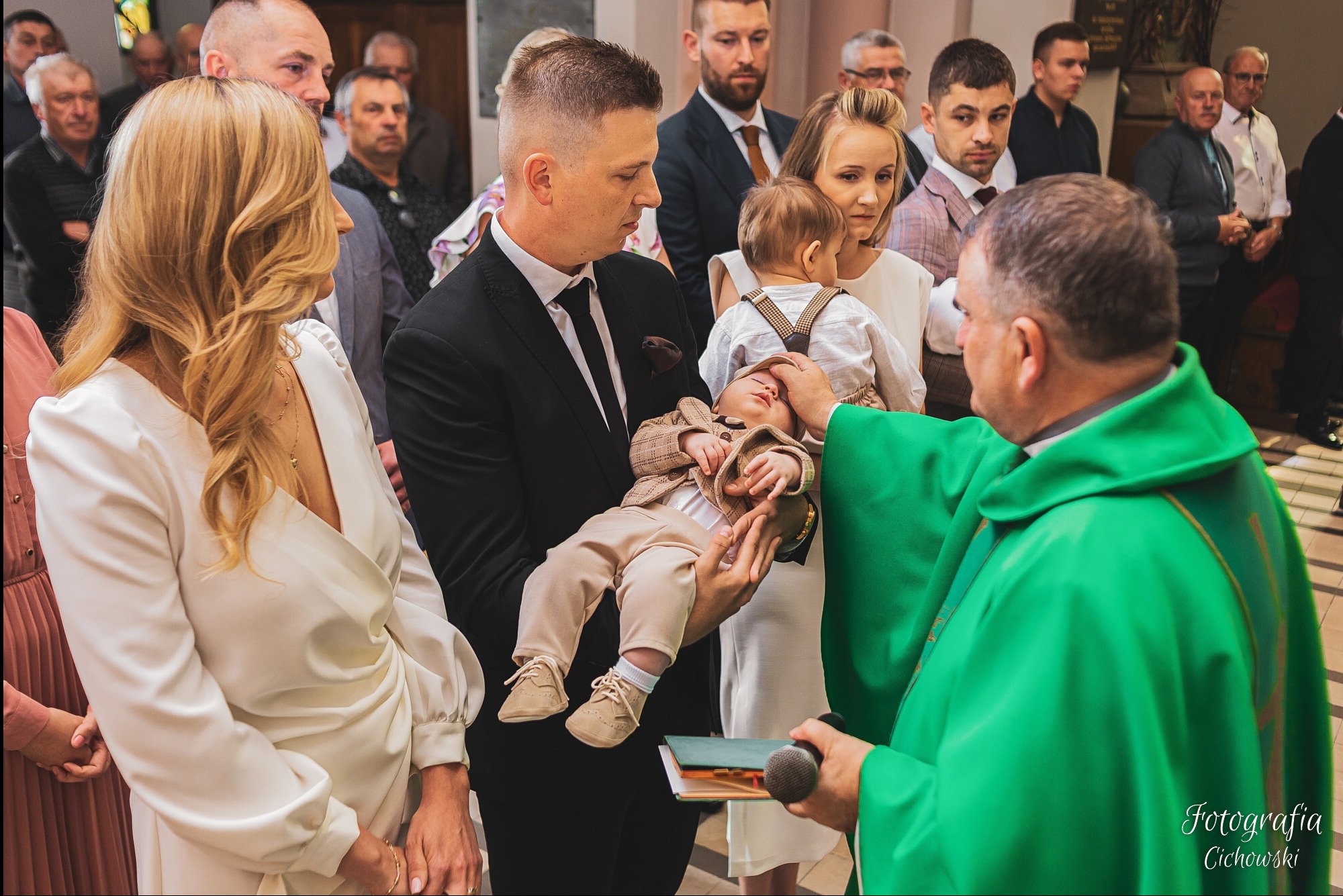 Ksiądz w zielonej szacie udziela sakramentu chrztu niemowlęciu ubranemu w beżowy garnitur, trzymanemu przez ojca w czarnym garniturze, w tle rodzina.