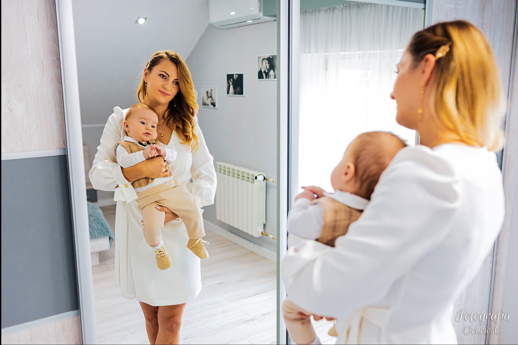 Elegancka mama w białej sukience trzymająca na rękach ubranego w brązową kamizelkę i beżowe spodnie niemowlaka, odbicie w lustrze szafy przesuwnej, w tle zdjęcia rodzinne na ścianie.