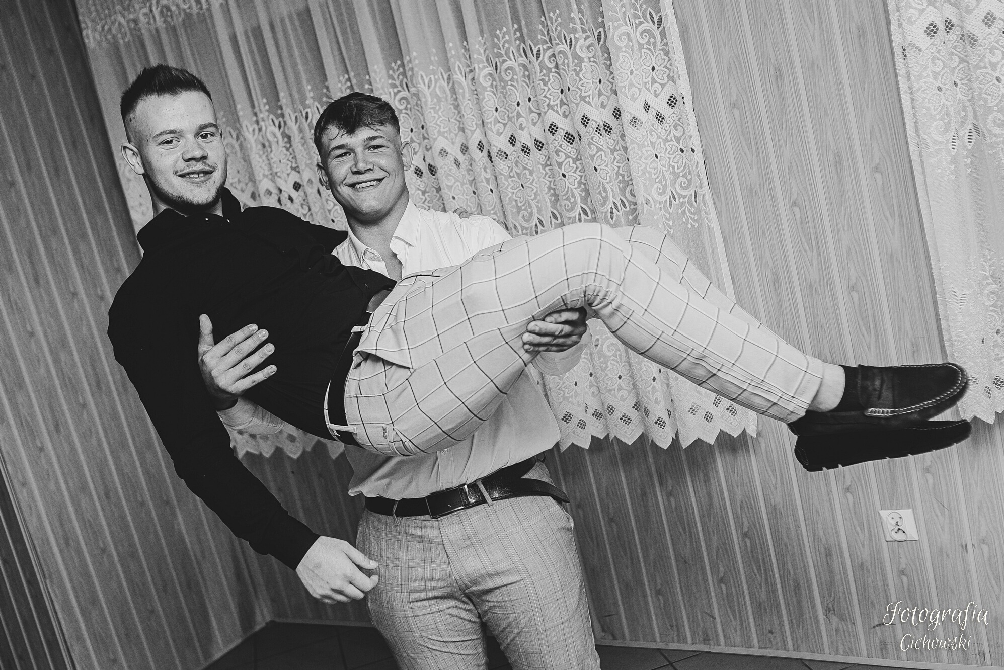 Czarno-białe zdjęcie dwóch uśmiechniętych mężczyzn, jeden niesie drugiego na rękach, w tle wzorzysta zasłona i boazeria.