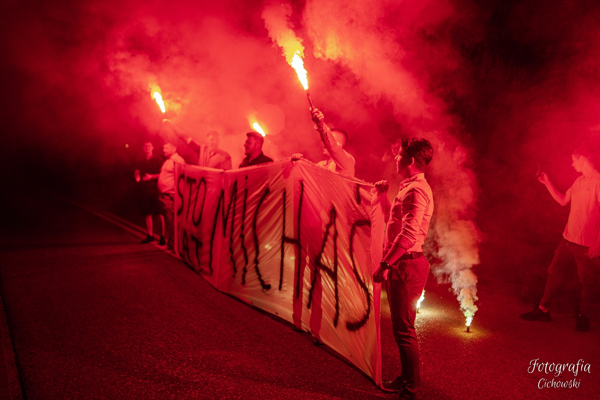Grupa osób trzymająca transparent z napisem 'STO MICHAŁ' podczas wydarzenia oświetlonego czerwonymi racami i dymem.