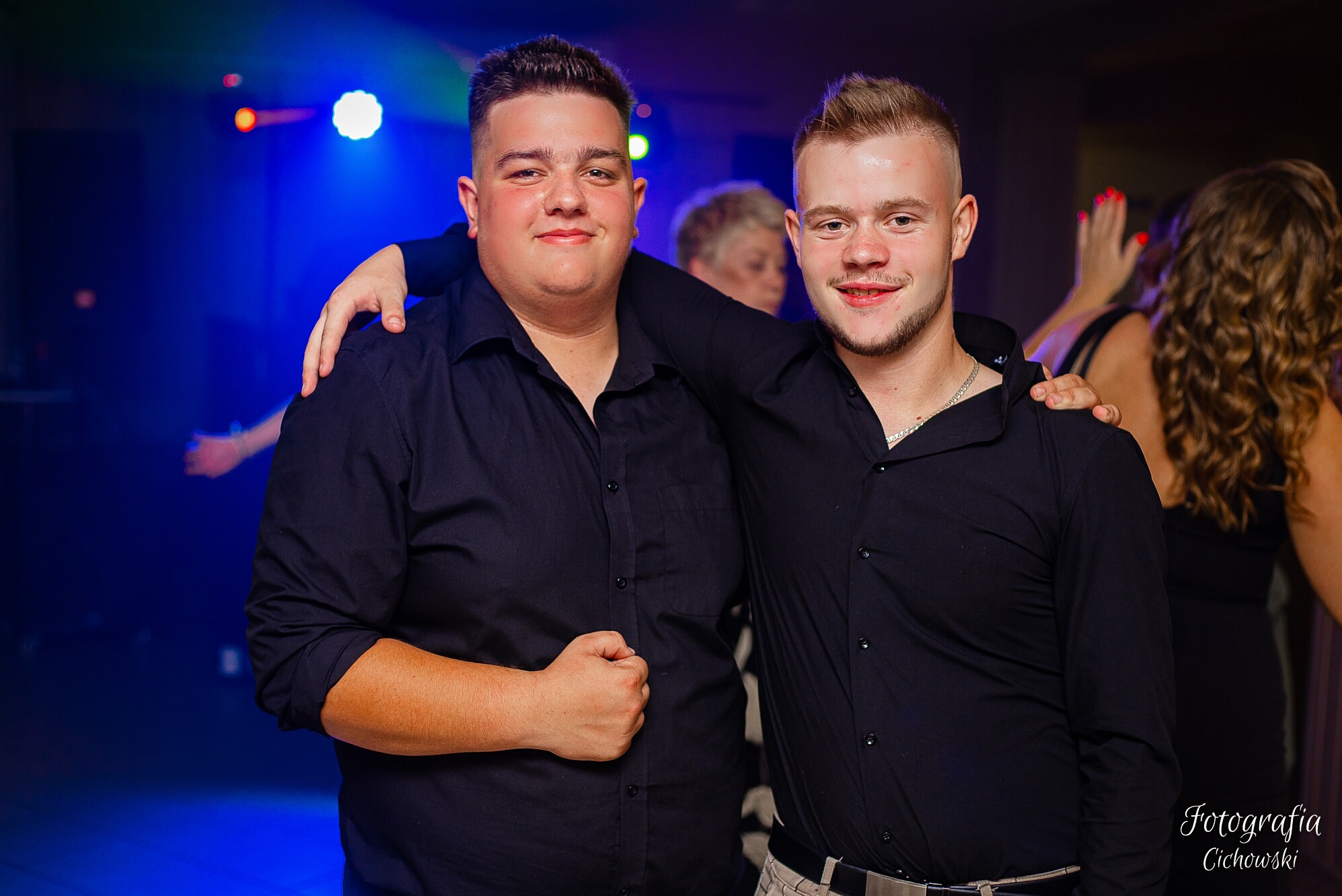Dwóch uśmiechniętych młodych mężczyzn w czarnych koszulach pozuje na tle sali z oświetleniem dyskotekowym podczas eventu.