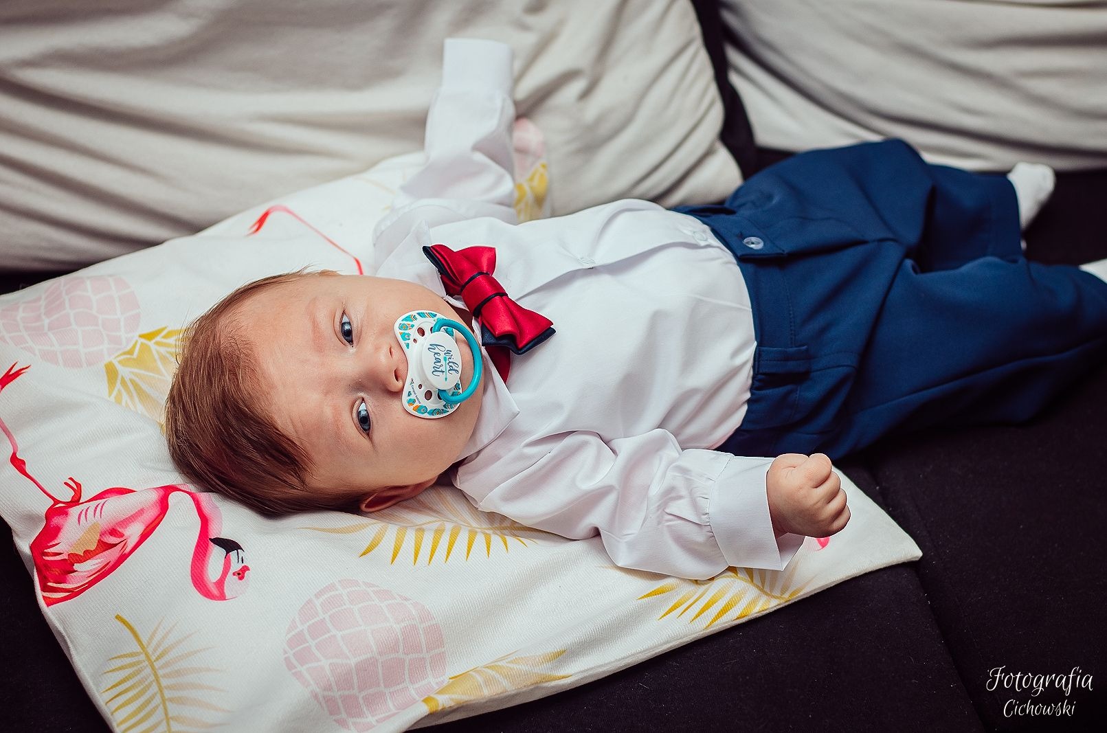 Uroczy niemowlak w eleganckim stroju leży na poduszce z motywem flamingów, ubrany w białą koszulę, granatowe spodnie i czerwoną muchę, z smoczkiem w ustach.