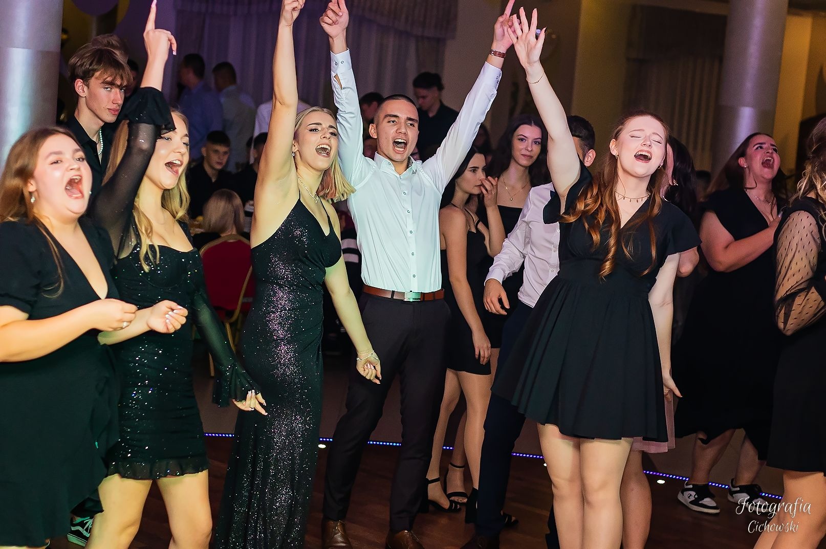 Grupa rozbawionych młodych ludzi w eleganckich strojach tańczy i śpiewa na parkiecie podczas imprezy, z uniesionymi rękami i radosnymi minami.