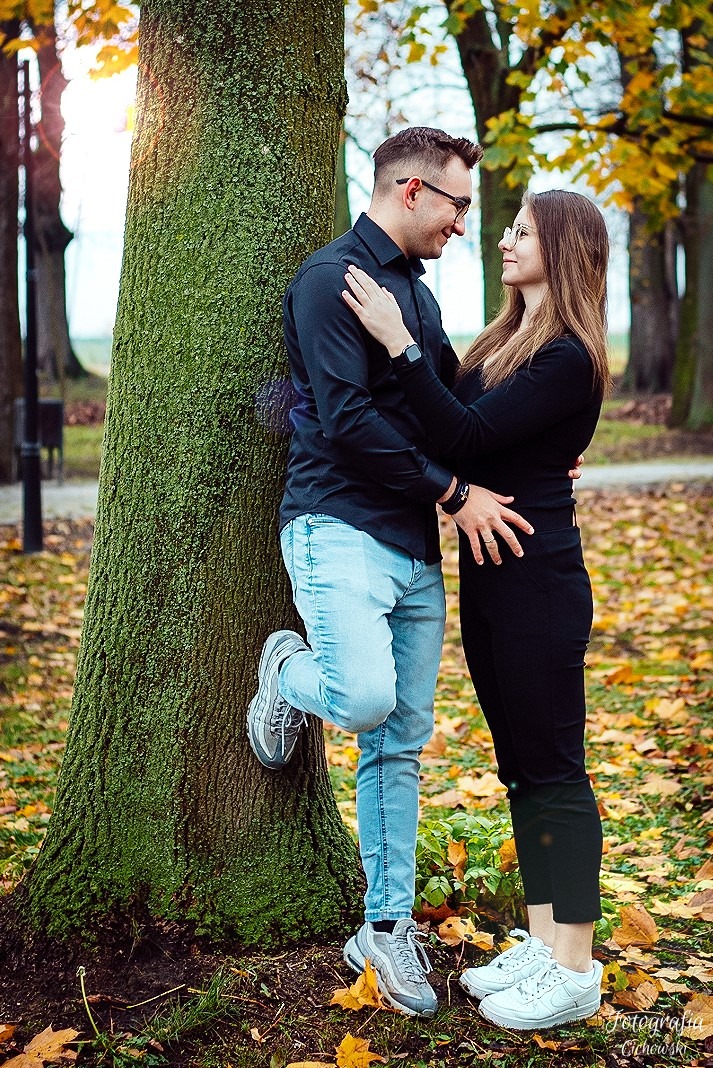 Para zakochanych oparta o drzewo w jesiennym parku, on w czarnej koszuli i jasnych jeansach, ona w czarnym stroju, oboje w sportowych butach i okularach.