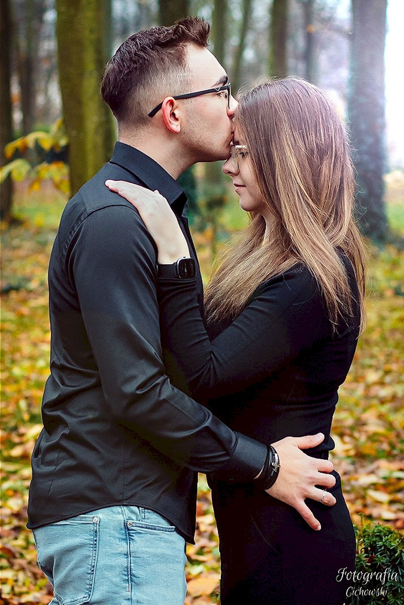 Mężczyzna w okularach całuje kobietę w czoło na tle jesiennego lasu, oboje ubrani na czarno, ona ma długie blond włosy.