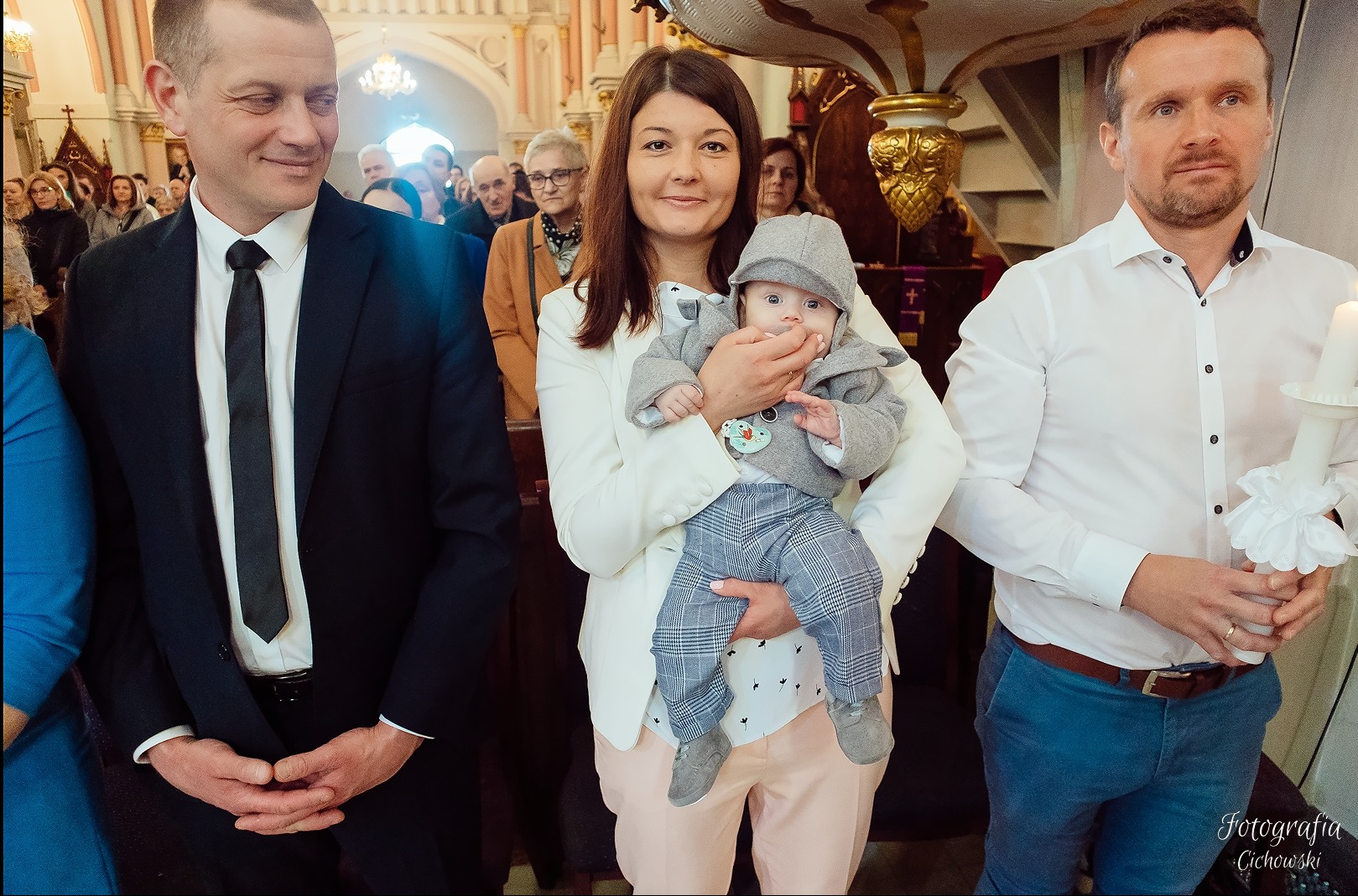 Uśmiechnięta matka trzyma na rękach elegancko ubranego chłopca w szarej czapeczce, obok mężczyźni w kościele, jeden trzyma świecę