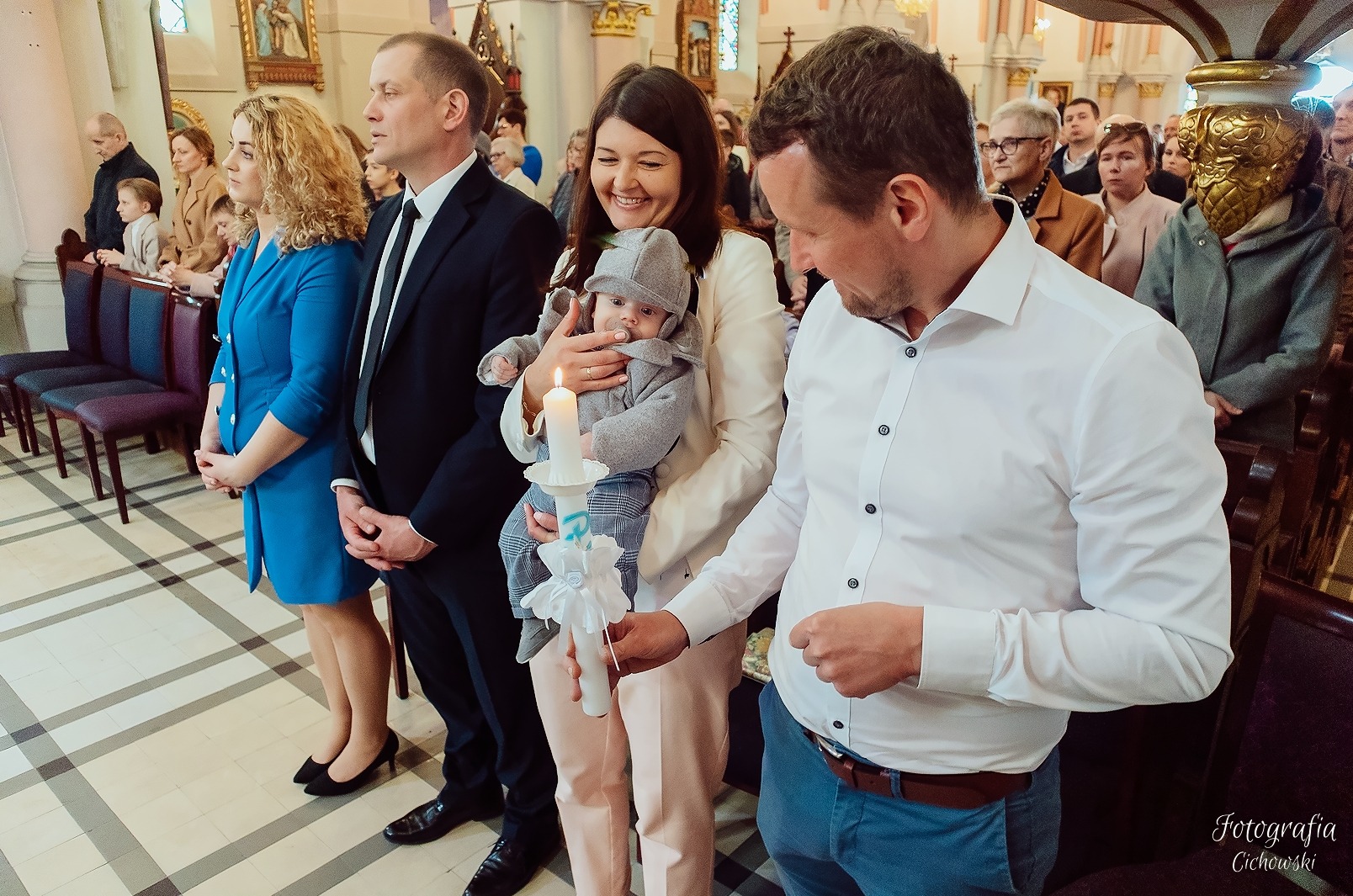 Rodzina podczas uroczystości w kościele, matka trzyma niemowlę w szarym ubranku i czapeczce, ojciec zapala świecę, w tle inni goście.