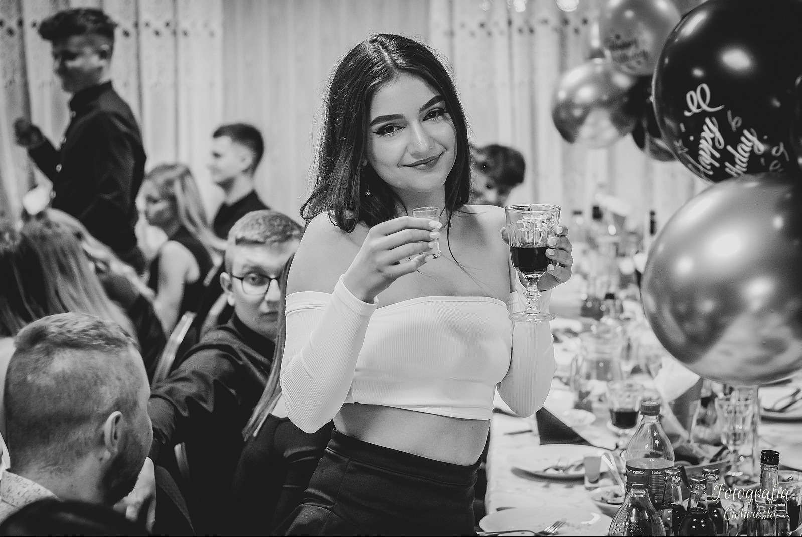 Uśmiechnięta brunetka w białej bluzce z odkrytymi ramionami, trzymająca kieliszek czerwonego wina i mały kieliszek wódki na przyjęciu z balonami w tle.