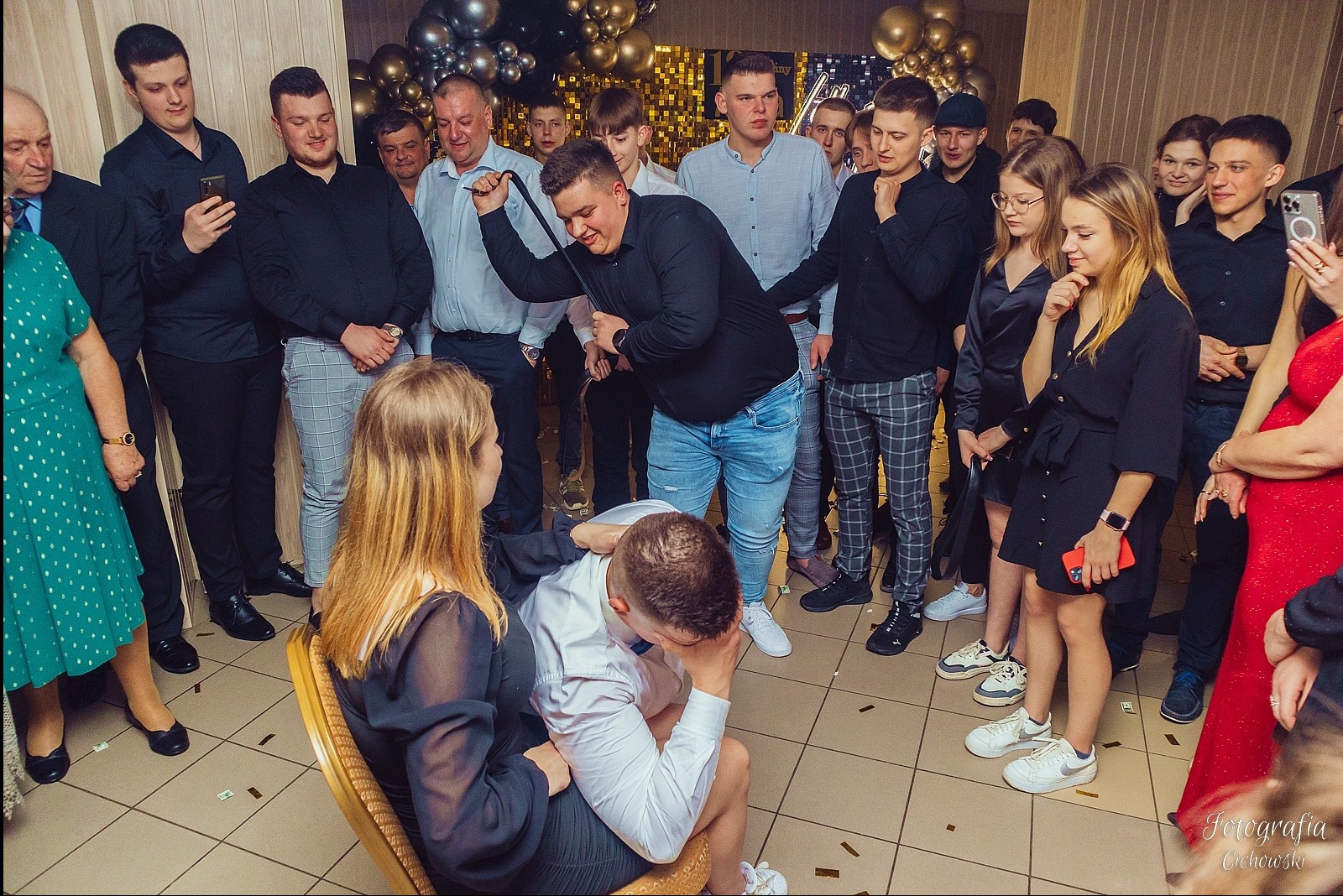 Gość weselny klęczy przed panną młodą podczas oczepin, otoczony grupą obserwujących osób, w tle dekoracja z balonów.