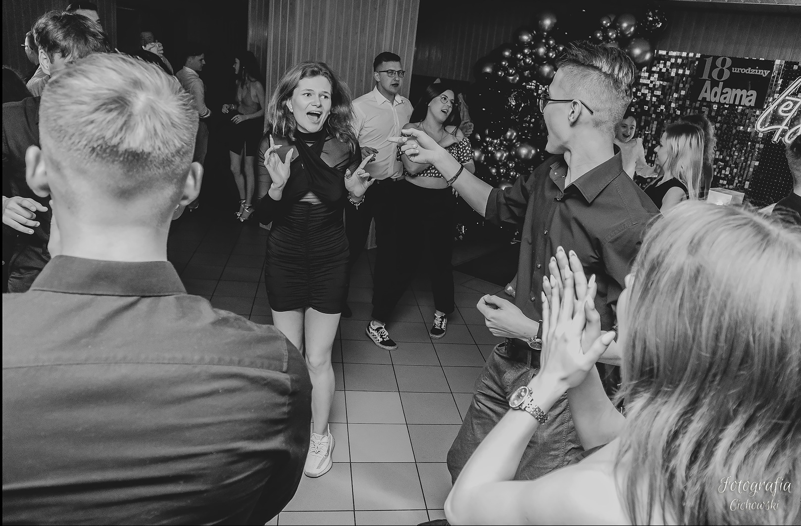 Czarno-białe zdjęcie tańczących osób na imprezie urodzinowej, z balonami i dekoracjami w tle, widać radość i energię uczestników.