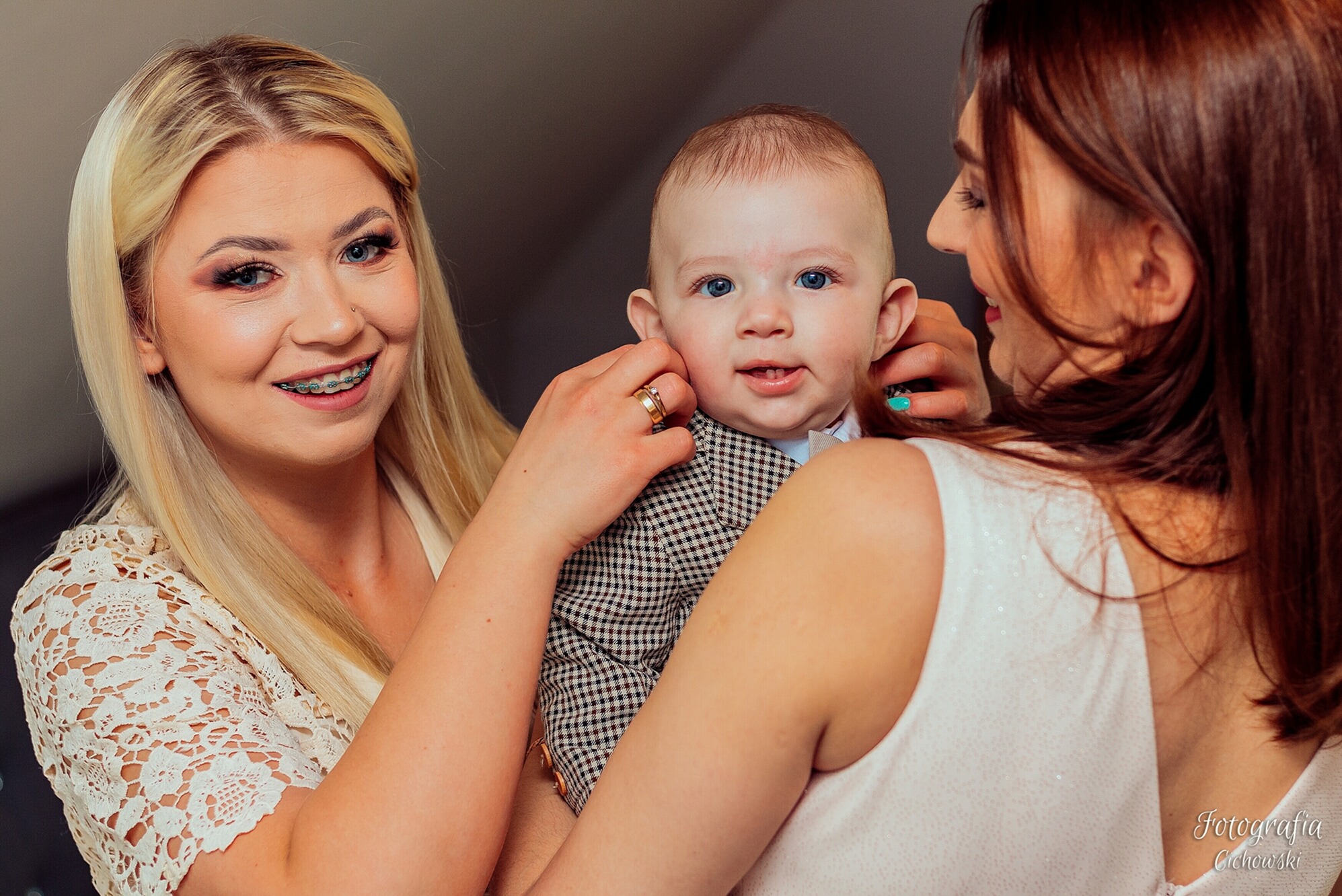 Dwie kobiety trzymające niemowlę w eleganckim ubranku; blondynka z aparatem ortodontycznym i pierścionkiem na palcu, uśmiechnięte dziecko z niebieskimi oczami.