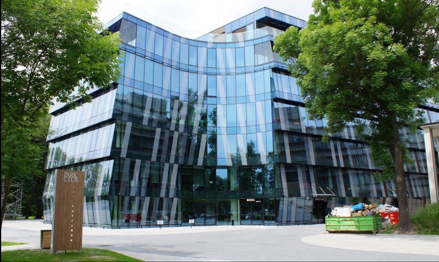 Nowoczesny budynek biurowo-apartamentowy z przeszkloną fasadą o nietypowym kształcie, otoczony zielenią. Widoczny znak 'Park Club' i kontener na śmieci.