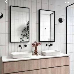Tomasz Jarosz - Nowoczesna łazienka z dwoma prostokątnymi umywalkami nablatowymi, czarnymi bateriami i lustrami w czarnych ramach na tle białych, pionowo ułożonych płytek. Na blacie stoi wazon z suszonymi kwiatami...