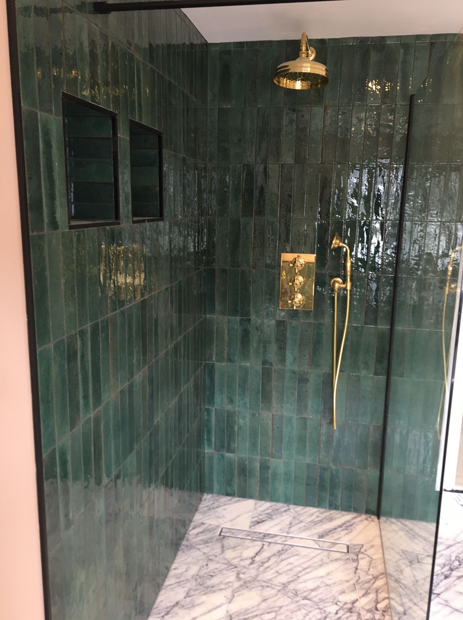 Nowoczesna kabina prysznicowa z zielonymi, błyszczącymi płytkami ułożonymi pionowo, złotą armaturą i marmurową posadzką z odpływem liniowym.