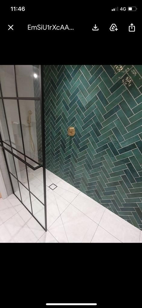 Kabina prysznicowa ze szklaną ścianką o czarnych profilach, zielone płytki ułożone w jodełkę na ścianie, biała podłoga z geometrycznym wzorem, złoty kran.