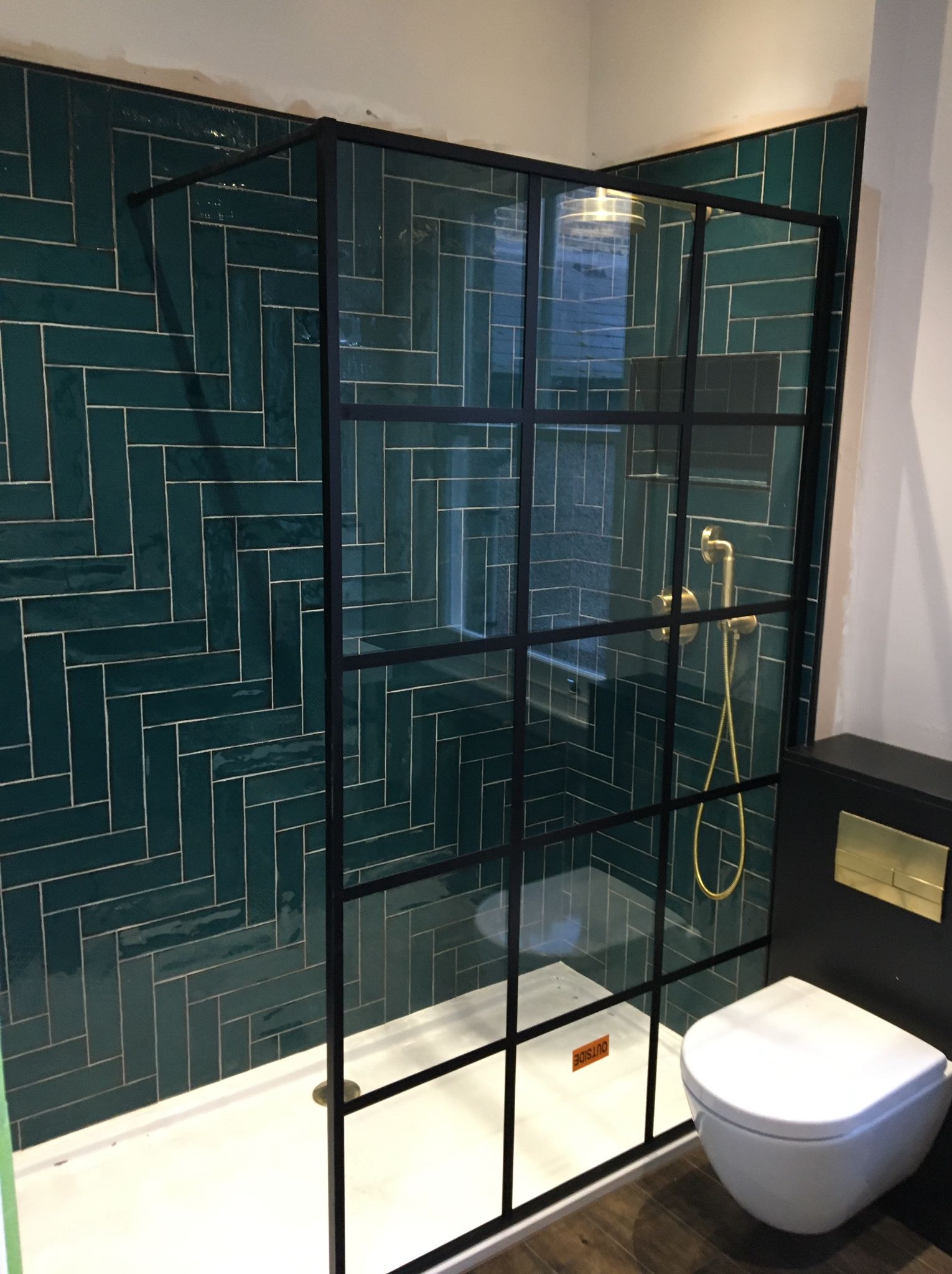 Nowoczesna łazienka z kabiną prysznicową typu walk-in z czarnymi profilami, zielone płytki ułożone w jodełkę na ścianie, biały brodzik i wisząca toaleta.