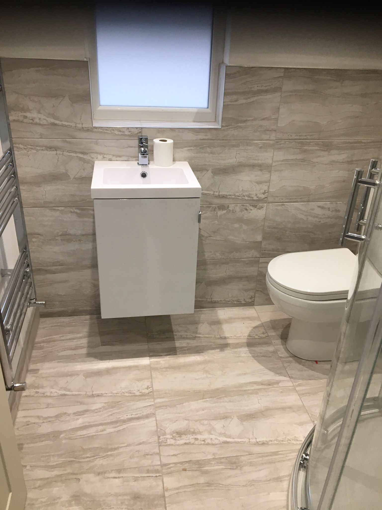 Nowoczesna łazienka wykończona w beżowych płytkach imitujących marmur, z podwieszaną szafką umywalkową, toaletą i grzejnikiem drabinkowym.