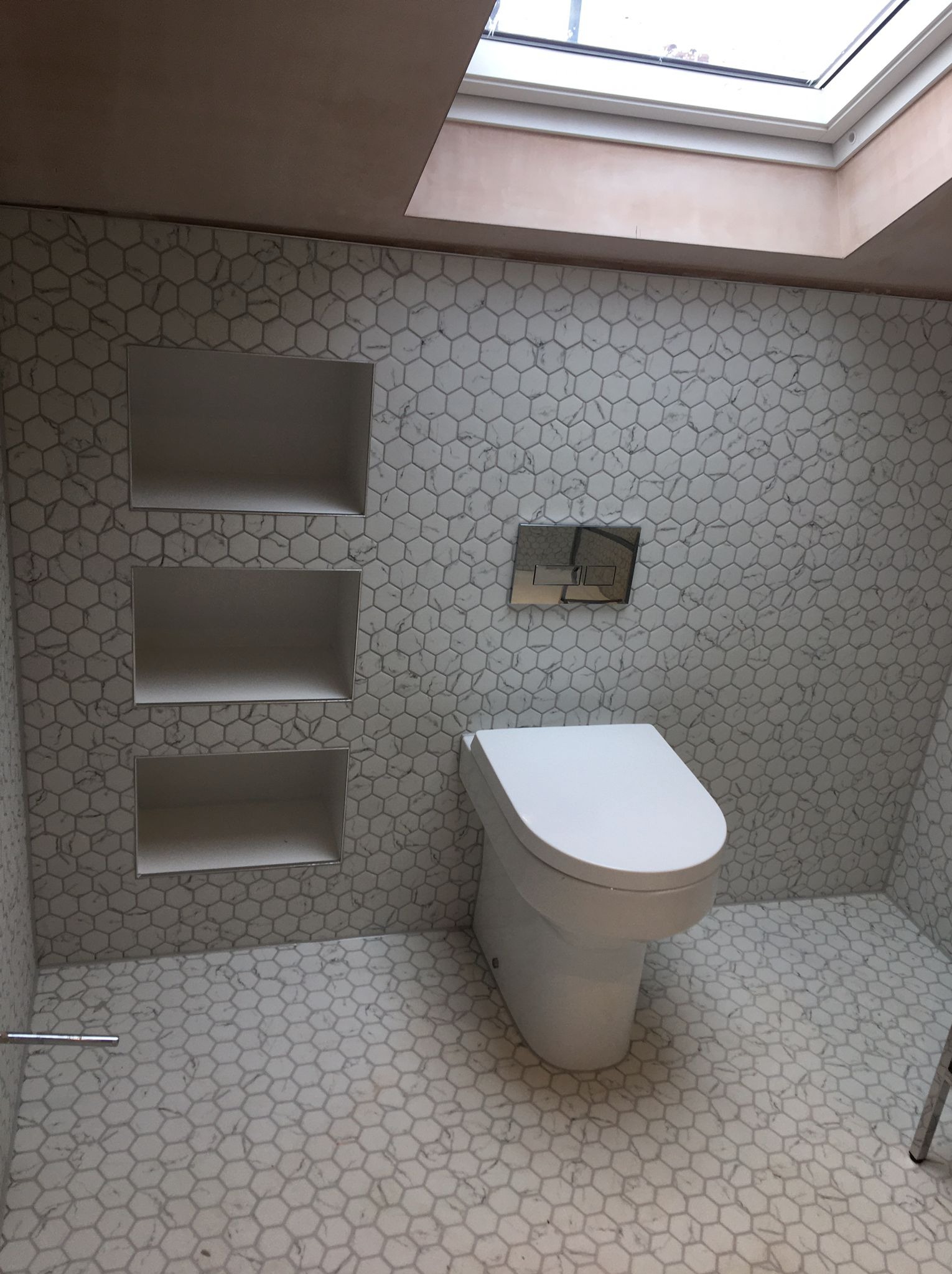 Wykończona łazienka z toaletą, heksagonalnymi płytkami podłogowymi i ściennymi w odcieniach bieli i szarości, wnękami w ścianie oraz oknem dachowym.